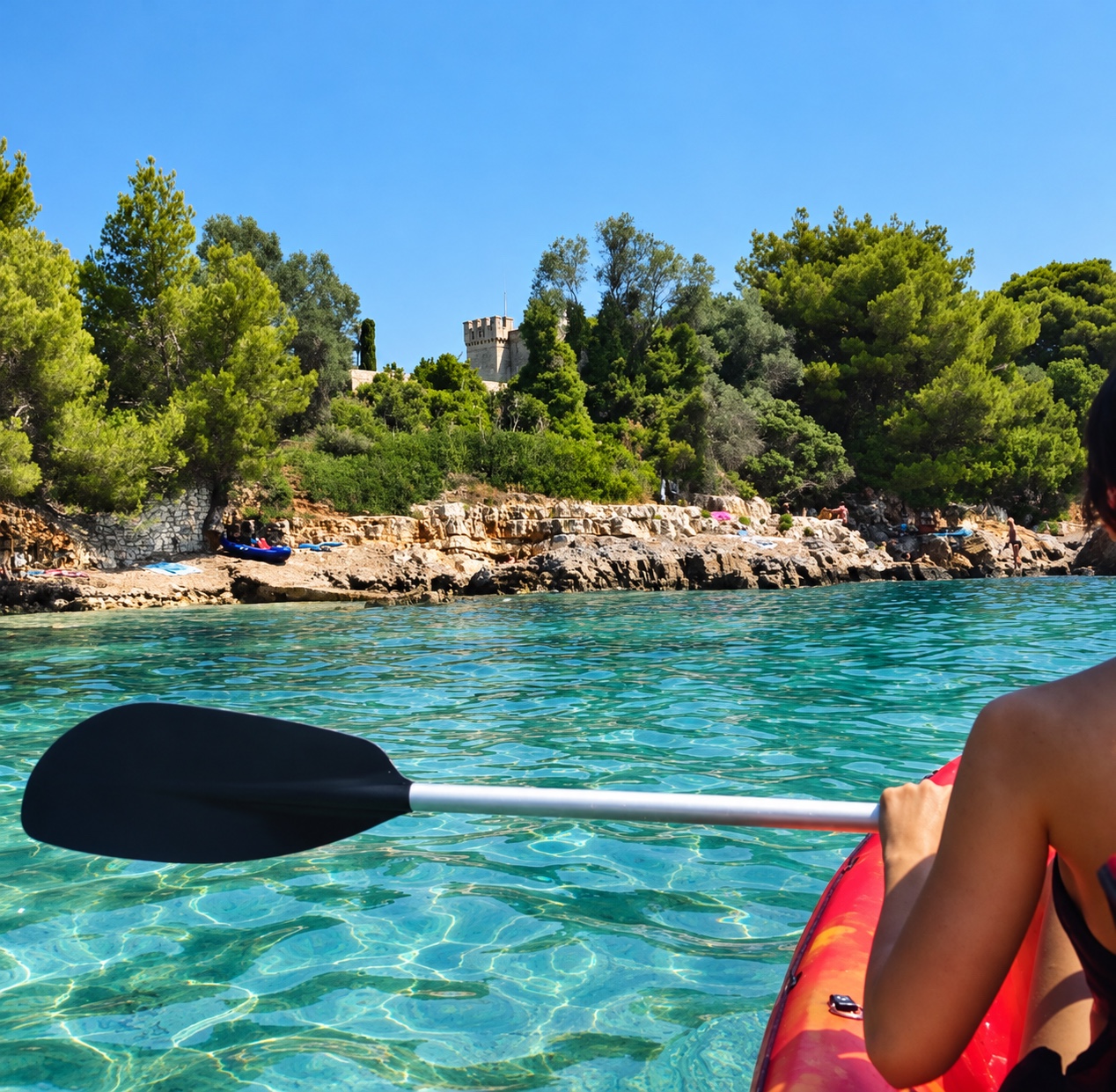 Une kayakiste longe un rivage boisé des îles de Lérins sur une eau turquoise.